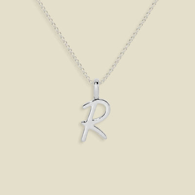 A silver initial necklace featuring an uppercase polished cursive letter 'R' pendant on a delicate silver chain, set against a solid light beige background.