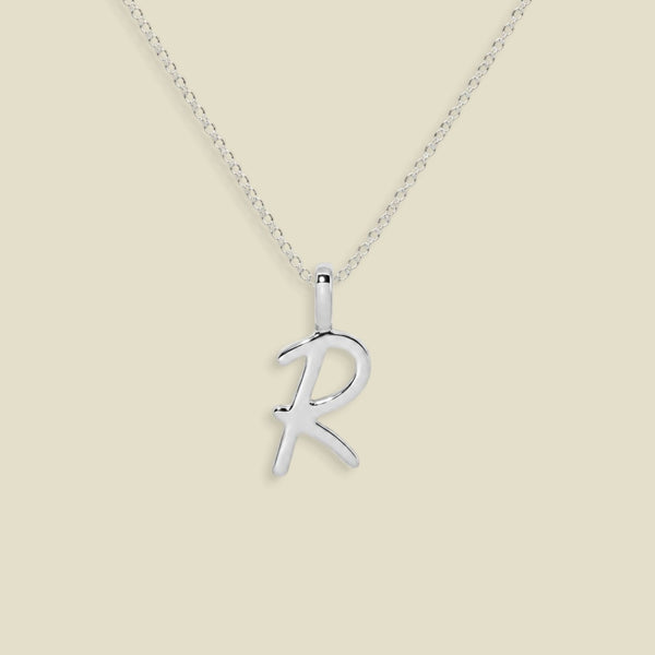 A silver initial necklace featuring an uppercase polished cursive letter 'R' pendant on a delicate silver chain, set against a solid light beige background.