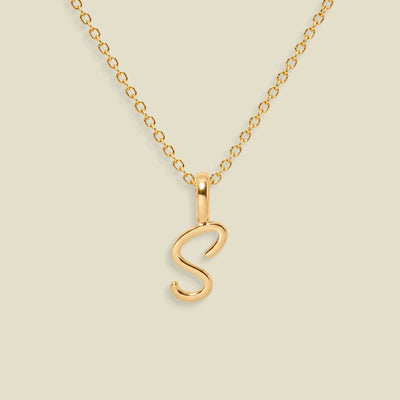 A gold initial necklace featuring a polished script letter 'S' pendant on a delicate gold chain, set against a solid light beige background.
