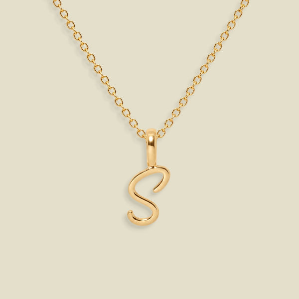 A gold initial necklace featuring a polished script letter 'S' pendant on a delicate gold chain, set against a solid light beige background.