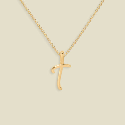 A gold initial pendant necklace featuring the uppercase letter 'T' in a cursive script, suspended from a delicate gold chain against a solid light beige background.
