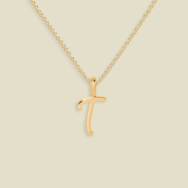 A gold initial pendant necklace featuring the uppercase letter 'T' in a cursive script, suspended from a delicate gold chain against a solid light beige background.