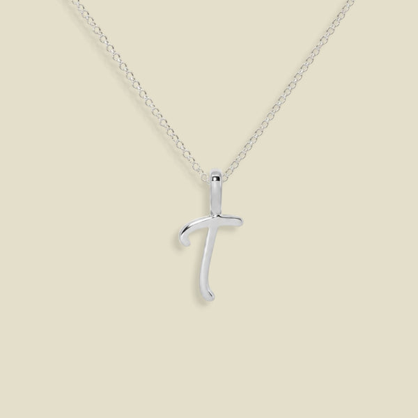 A silver initial pendant necklace featuring the uppercase letter 'T' on a delicate silver chain, set against a solid beige background.