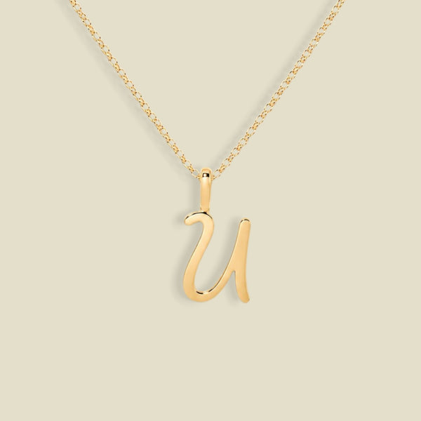 A gold initial necklace featuring a cursive uppercase letter 'U' pendant on a delicate gold chain, set against a solid light beige background.