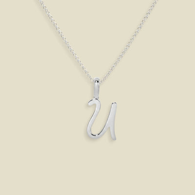 A silver initial necklace featuring an uppercase cursive letter 'U' pendant on a delicate silver chain, set against a solid light beige background.