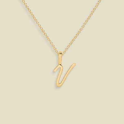A gold initial necklace featuring a polished uppercase script letter 'V' pendant on a delicate gold chain, set against a solid light beige background.
