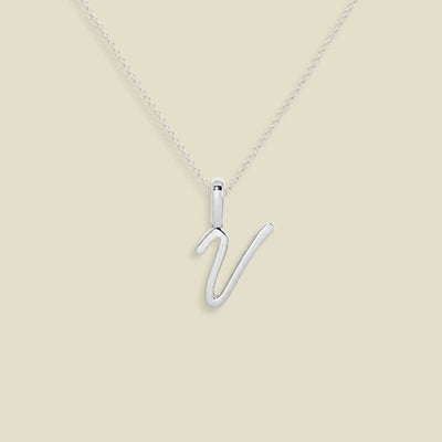 A silver initial necklace featuring a polished uppercase script letter 'V' pendant on a delicate silver chain, set against a solid beige background.