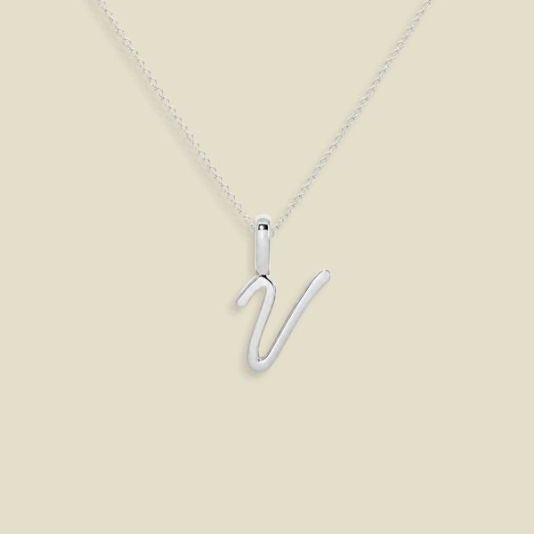 A silver initial necklace featuring a polished uppercase script letter 'V' pendant on a delicate silver chain, set against a solid beige background.