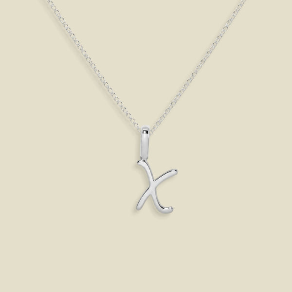 A silver initial necklace featuring a polished cursive uppercase letter 'X' pendant on a delicate link chain, set against a solid light beige background.