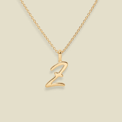 A gold initial necklace featuring a polished cursive letter 'Z' pendant on a delicate gold chain, set against a solid light beige background.