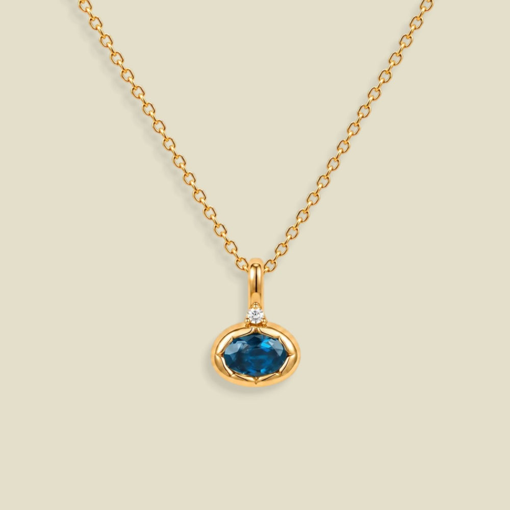 A gold chain necklace with a pendant featuring an oval dark blue-green gemstone in a gold bezel setting, accented by a small clear stone on the top, set against a solid light beige background.