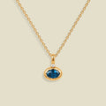 A gold chain necklace with a pendant featuring an oval dark blue-green gemstone in a gold bezel setting, accented by a small clear stone on the top, set against a solid light beige background.