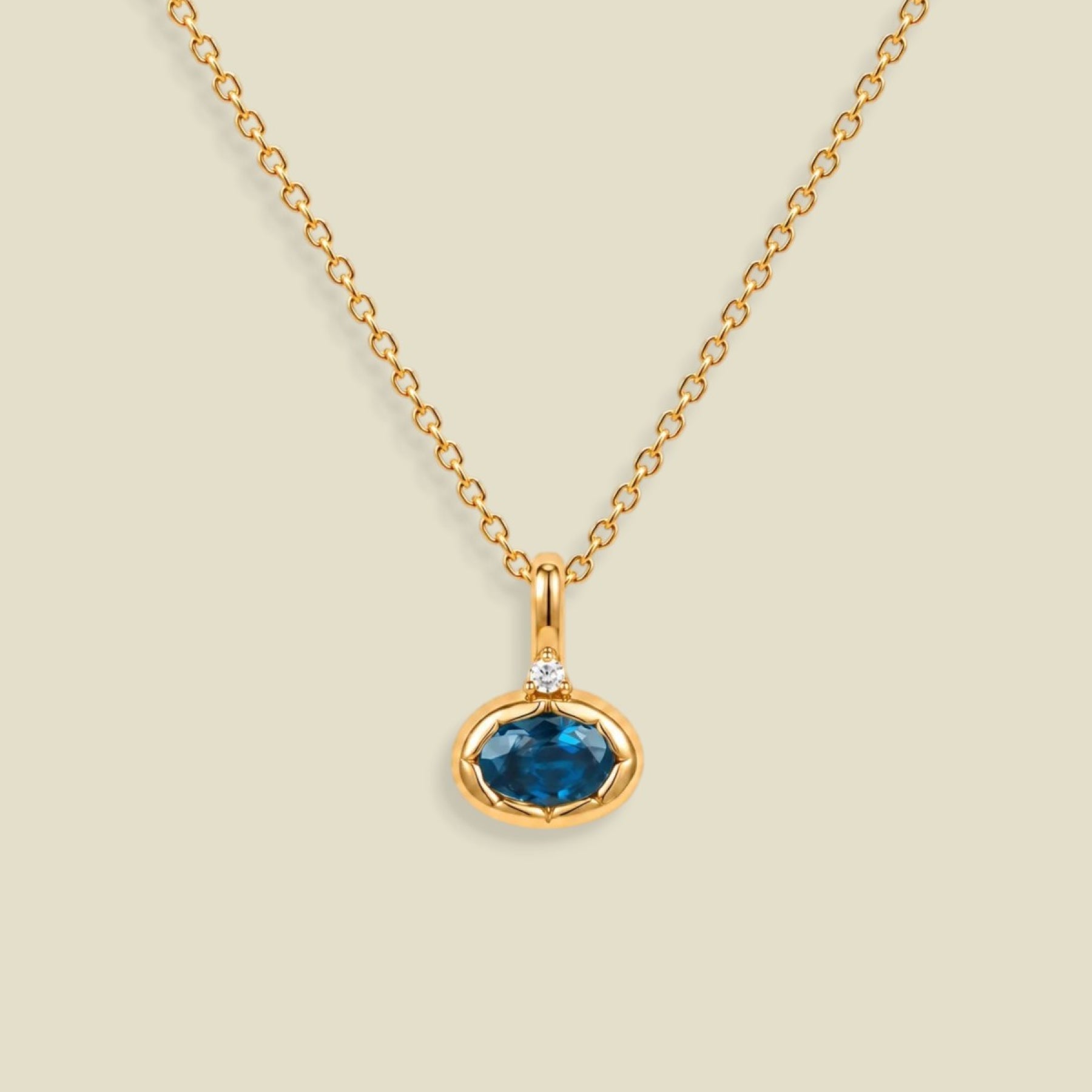 A gold chain necklace with a pendant featuring an oval dark blue-green gemstone in a gold bezel setting, accented by a small clear stone on the top, set against a solid light beige background.