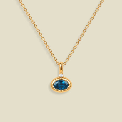 A gold chain necklace with a pendant featuring an oval dark blue-green gemstone in a gold bezel setting, accented by a small clear stone on the top, set against a solid light beige background.