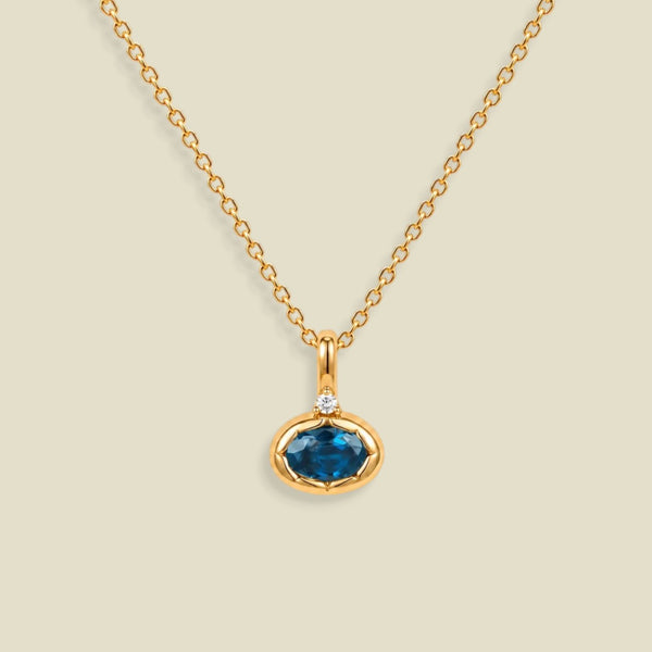 A gold chain necklace with a pendant featuring an oval dark blue-green gemstone in a gold bezel setting, accented by a small clear stone on the top, set against a solid light beige background.