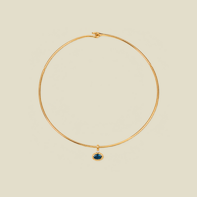 A thin gold wire hoop necklace with a small oval blue-green gemstone pendant, displayed on a solid light beige background.