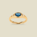 A gold signet ring with a blue-green center oval bezel-set gemstone, and two small clear accent stones on either side, set against a solid beige background.