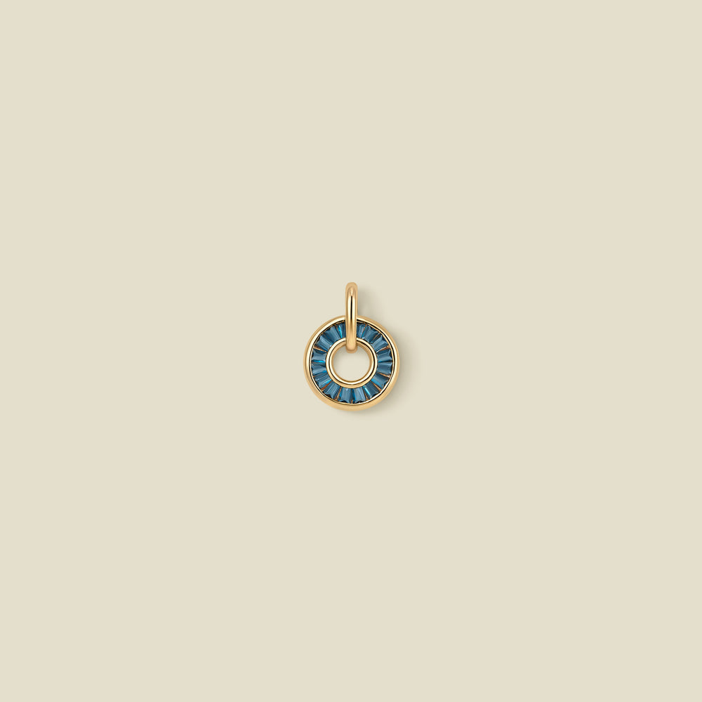 A gold circular pendant featuring blue-green baguette-cut stones around a hollow center, displayed against a solid light beige background.