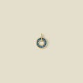 A gold circular pendant featuring blue-green baguette-cut stones around a hollow center, displayed against a solid light beige background.