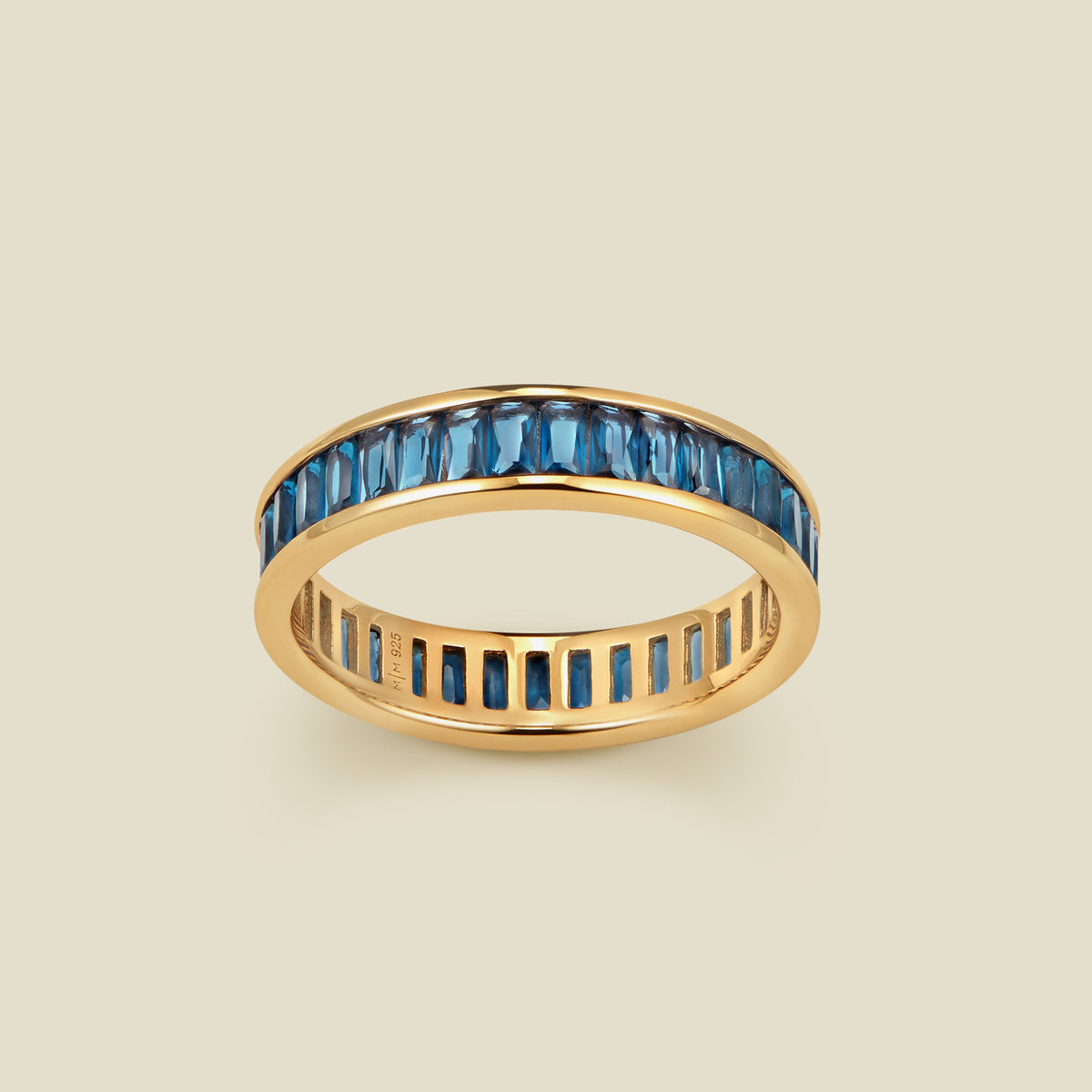 A gold eternity ring featuring a continuous channel setting of baguette-cut blue-green gemstones, shown on a solid light beige background.