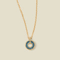 A gold chain necklace with a circular pendant featuring blue-green baguette-cut stones around a hollow center, displayed against a solid light beige background.