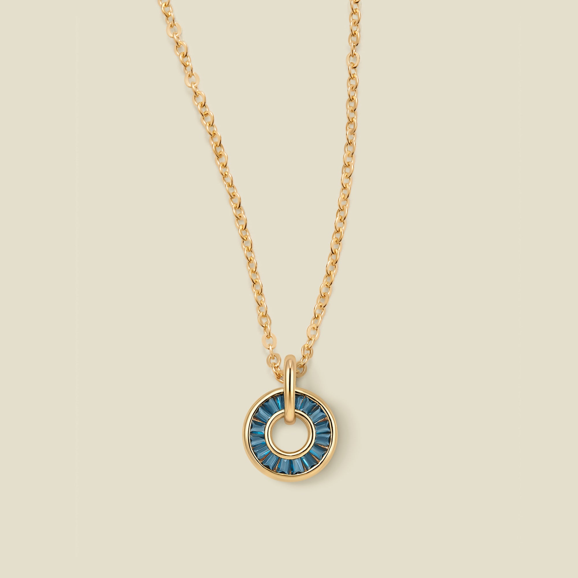 A gold chain necklace with a circular pendant featuring blue-green baguette-cut stones around a hollow center, displayed against a solid light beige background.