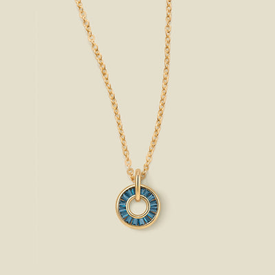 A gold chain necklace with a circular pendant featuring blue-green baguette-cut stones around a hollow center, displayed against a solid light beige background.