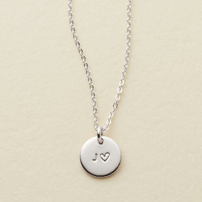 A silver chain necklace with a round disc pendant engraved with the letter 'J' and a heart next to it, displayed on a light beige textured background.