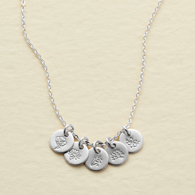 A silver chain necklace featuring five small round disc charms with engraved floral designs, displayed on a textured light beige background.