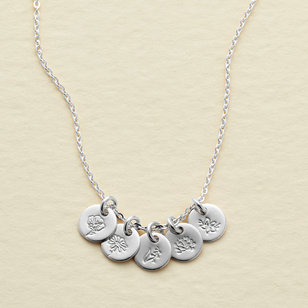 A silver chain necklace featuring five small round disc charms with engraved floral designs, displayed on a textured light beige background.