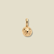 A small gold soccer ball charm pendant with a textured pattern, shown on a solid light beige background.