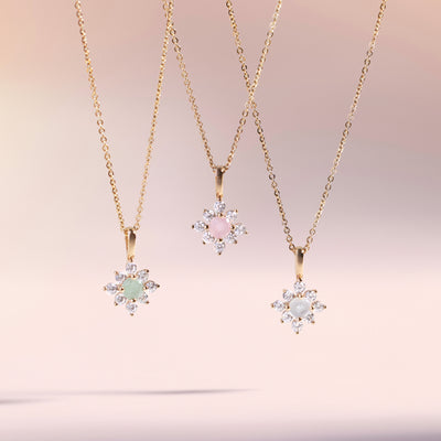 Three gold chains with a green, pink and white stone pendant suspended against a light pink backdrop. 