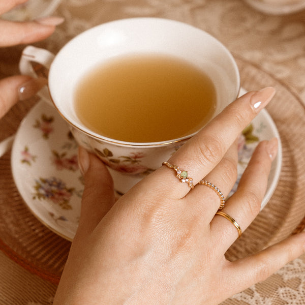 Woman holding a floral teacup, wearing delicate gold stacked rings.