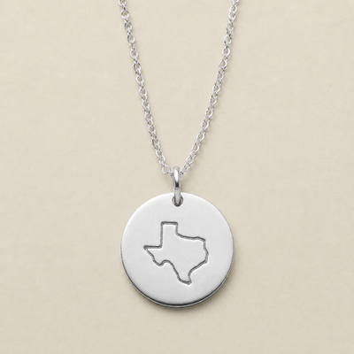 A silver round disc pendant necklace featuring a cutout outline of Texas, suspended from a delicate silver chain against a solid beige background.