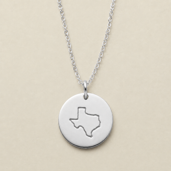 A silver round disc pendant necklace featuring a cutout outline of Texas, suspended from a delicate silver chain against a solid beige background.