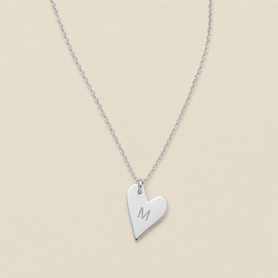 A silver heart-shaped pendant necklace engraved with the letter 'M', displayed on a delicate silver chain against a solid beige background.