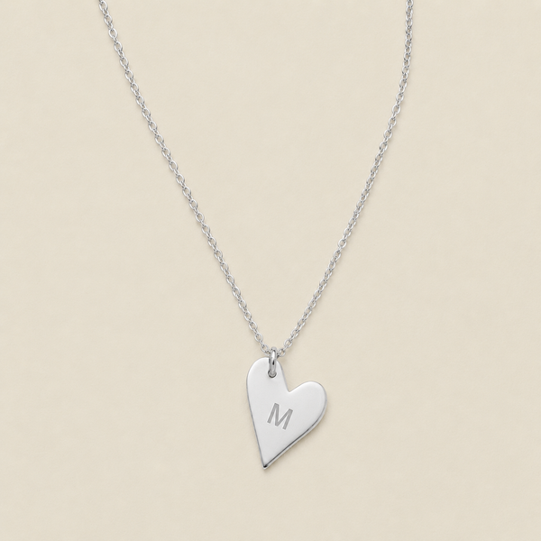 A silver heart-shaped pendant necklace engraved with the letter 'M', displayed on a delicate silver chain against a solid beige background.