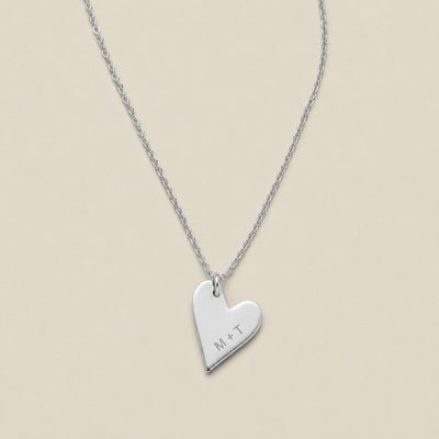 A silver heart-shaped pendant necklace engraved with the initials 'M+T', suspended from a delicate silver chain against a solid beige background.