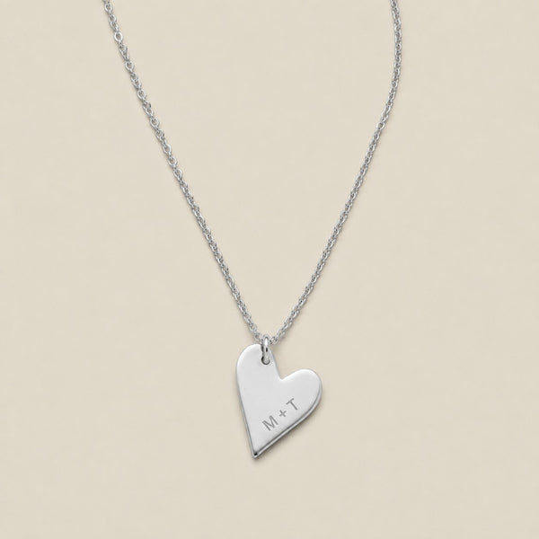 A silver heart-shaped pendant necklace engraved with the initials 'M+T', suspended from a delicate silver chain against a solid beige background.