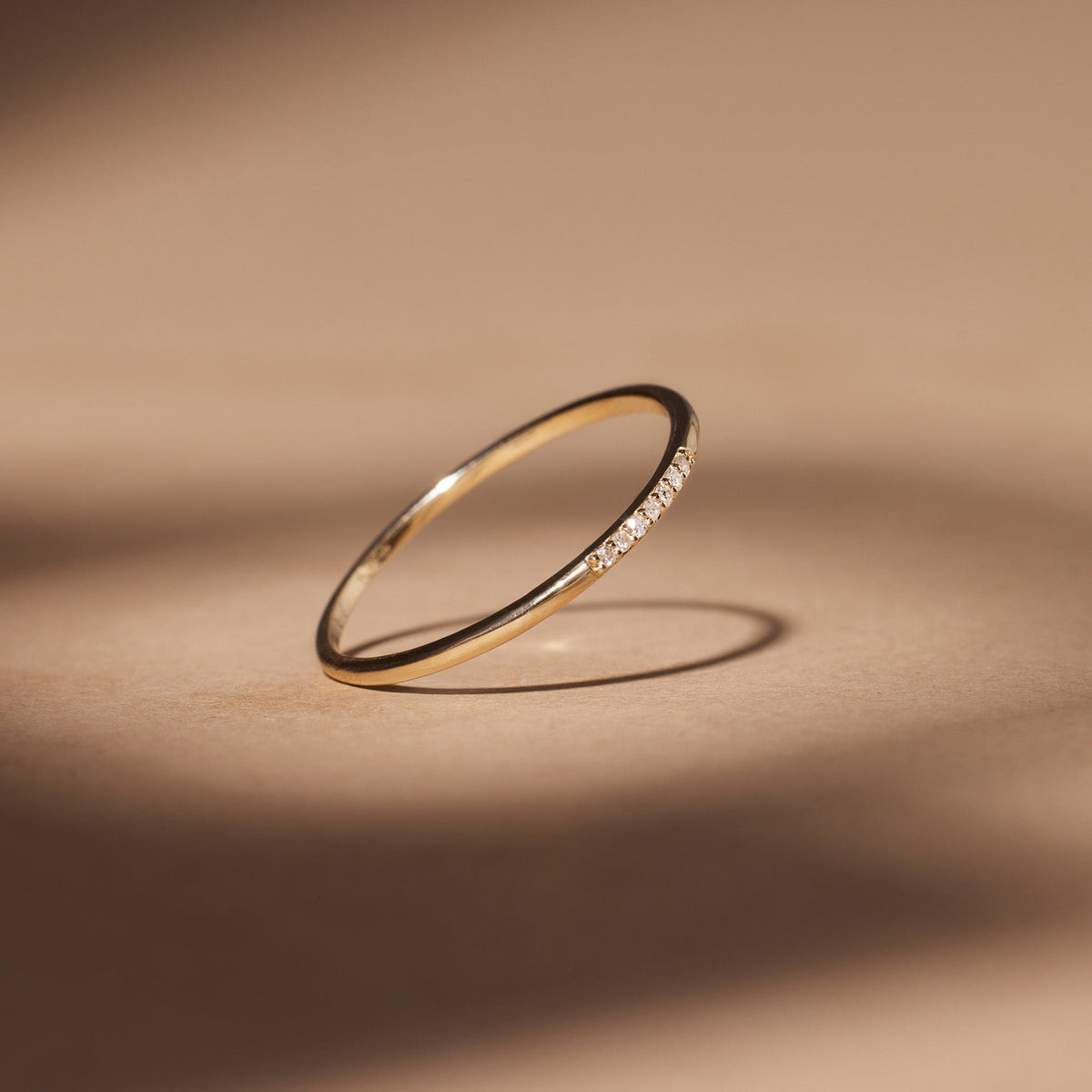 A polished gold thin band ring with 7 diamonds side by side centered in the front, pictured at an angle against a light brown background with moody shadows.