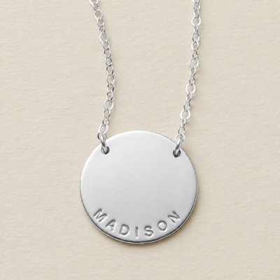 A silver round disc pendant necklace engraved with the name 'MADISON' at the bottom, hanging from a delicate silver chain against a textured beige background.