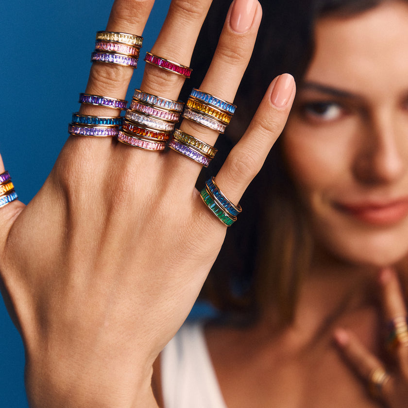 A close-up lifestyle shot of a hand wearing multiple colorful ring sets, with a woman's face softly blurred in the background against a solid blue backdrop.