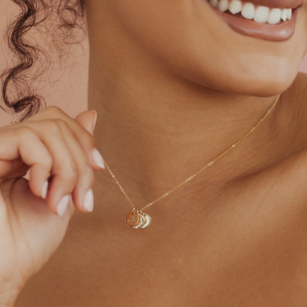 A close-up lifestyle shot of a model wearing a delicate gold chain necklace with three small stacked round charms, one of which features an engraved daisy design.