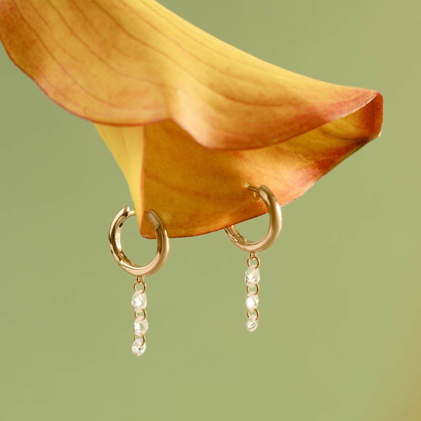 Two gold hoop earrings with three small clear crystals hanging vertically, set in a flower petal against a light green backdrop.