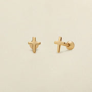A pair of gold cross stud earrings shown on a solid light beige background, with one earring facing forward and the other angled to show the post and backing.