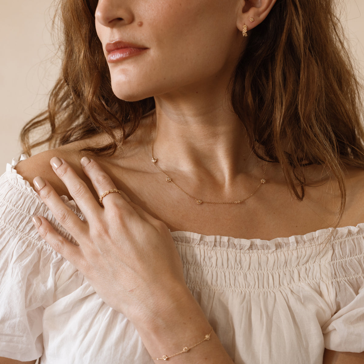 A close-up lifestyle shot of a woman wearing delicate gold jewelry, including a gold chain necklace with small stationary flower charms, a gold ring, and a gold bracelet, paired with a white off-the-shoulder top.