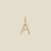 A gold letter 'A' pendant charm embellished with small clear round gemstones, centered on a solid light beige background.