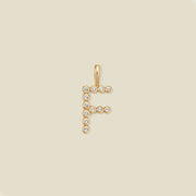 A gold letter 'F' pendant charm embellished with small clear round gemstones, centered on a solid light beige background.