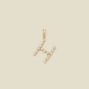 A gold letter 'H' pendant charm embellished with small clear gemstones, set against a solid light beige background.