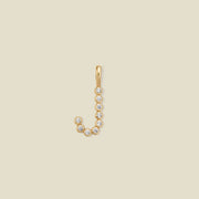 A gold letter 'J' initial pendant charm made up of clear round-cut bezel set gemstones, set against a solid light beige background.